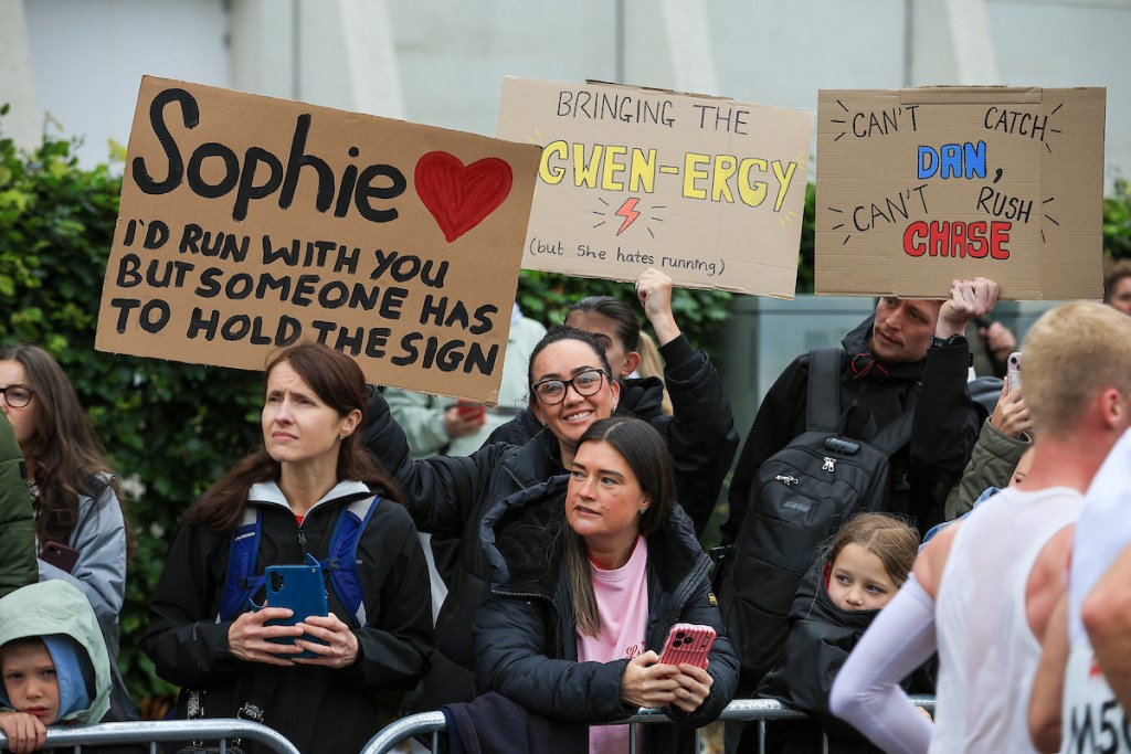 Spectators holding humorous handmade cardboard signs at the roadside.