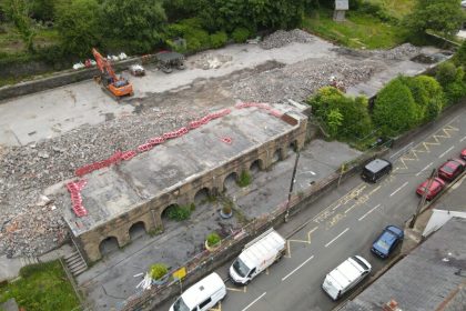 Aerial view of the part-demolished former Godre'rgraig Primary School, with an excavator working on cleared ground and rubble beside the remaining arched stone wall along Graig Road.