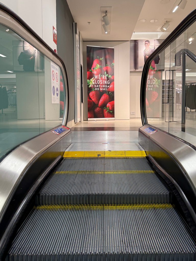 View up a stationary escalator inside M&S Swansea towards a banner reading "We're closing on Saturday 30 May"