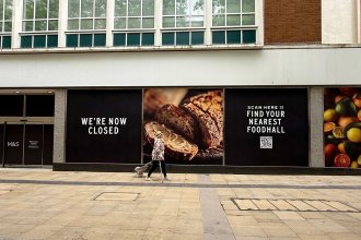 Boarded and blacked-out M&S store windows on Oxford Street, Swansea, with a lone shopper passing by
