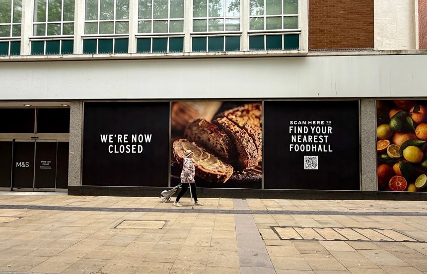 Boarded and blacked-out M&S store windows on Oxford Street, Swansea, with a lone shopper passing by