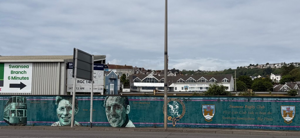 A long painted mural on the ground's perimeter wall depicting Swansea sporting figures, a vintage tram and club crests, with the words "Swansea Rugby Club, first ever club side to beat the Southern hemisphere 'Big Three'."