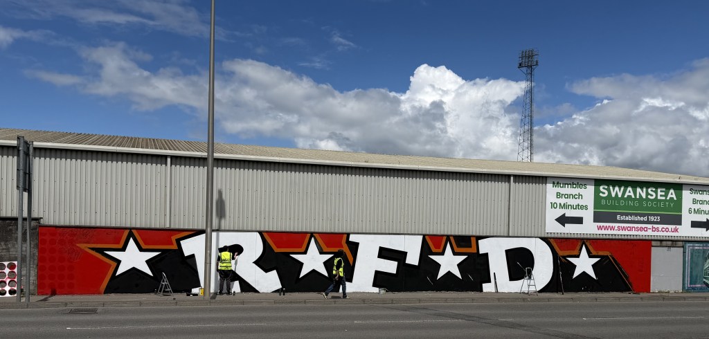 Two painters in hi-vis vests working on giant RFD graffiti letters with stars on the wall of St Helen's stadium under a blue sky