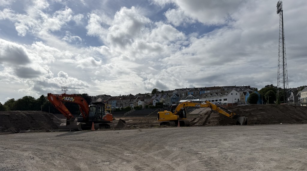 Two excavators working on the stripped earth of the former St Helen's pitch, with terraced houses and a floodlight pylon behind