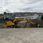 Yellow JCB excavator moving earth inside St Helen's stadium, with old terracing and the Swansea Cricket and Football Club building in the background
