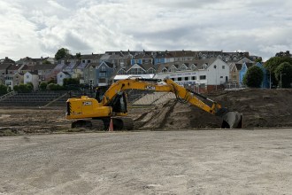 Yellow JCB excavator moving earth inside St Helen's stadium, with old terracing and the Swansea Cricket and Football Club building in the background
