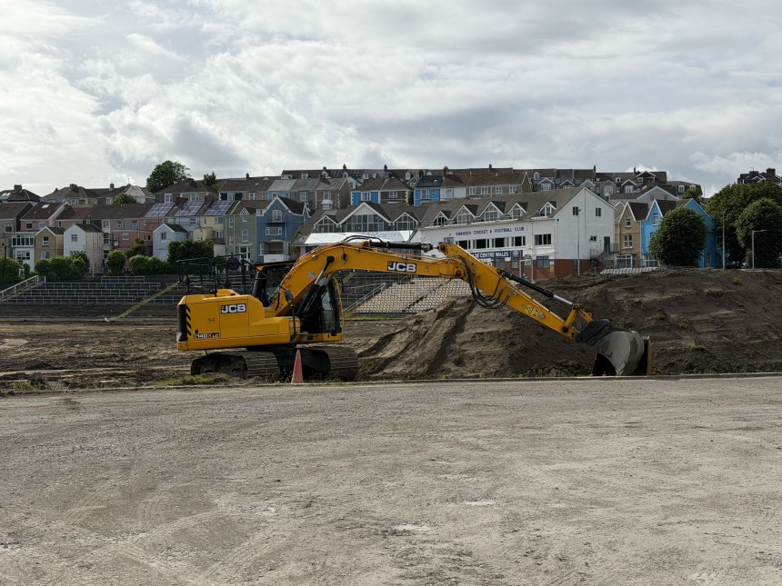Yellow JCB excavator moving earth inside St Helen's stadium, with old terracing and the Swansea Cricket and Football Club building in the background