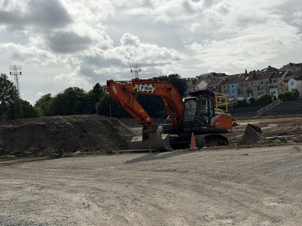 Orange hired excavator on bare earth inside St Helen's stadium with two floodlight pylons against a cloudy sky
