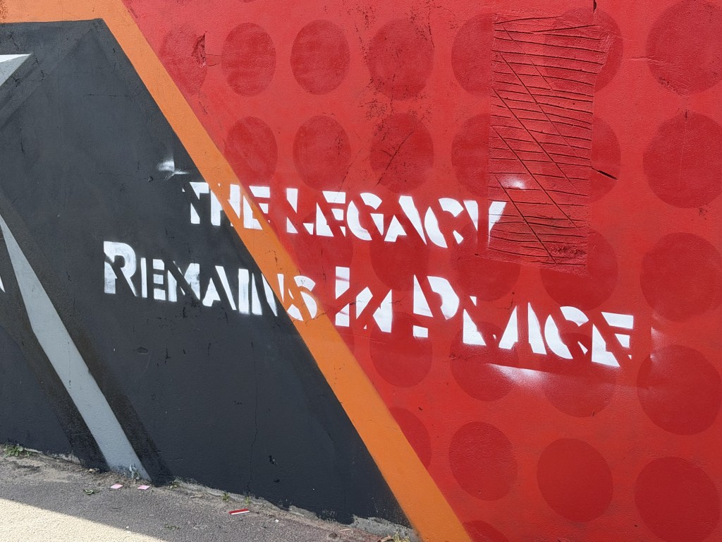 Close-up of white stencilled words The Legacy Remains in Place painted across red and black sections of the mural
