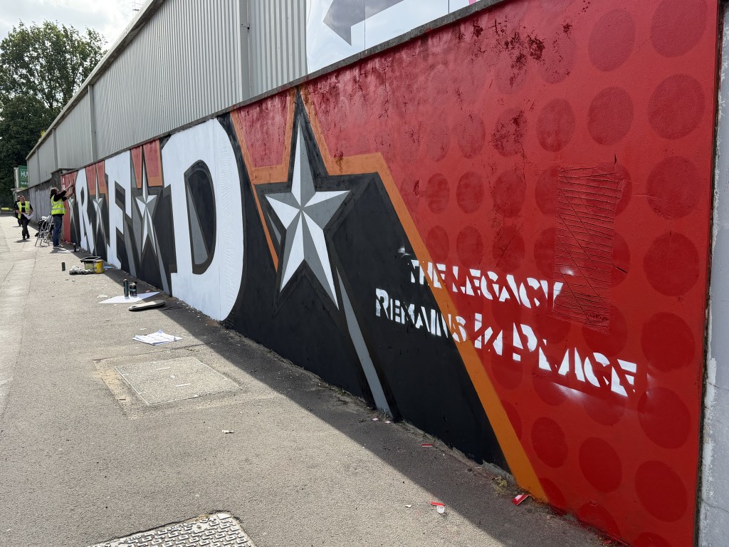 Angled view along the St Helen's stadium wall showing the giant letter D, a white star and the stencilled words The Legacy Remains in Place