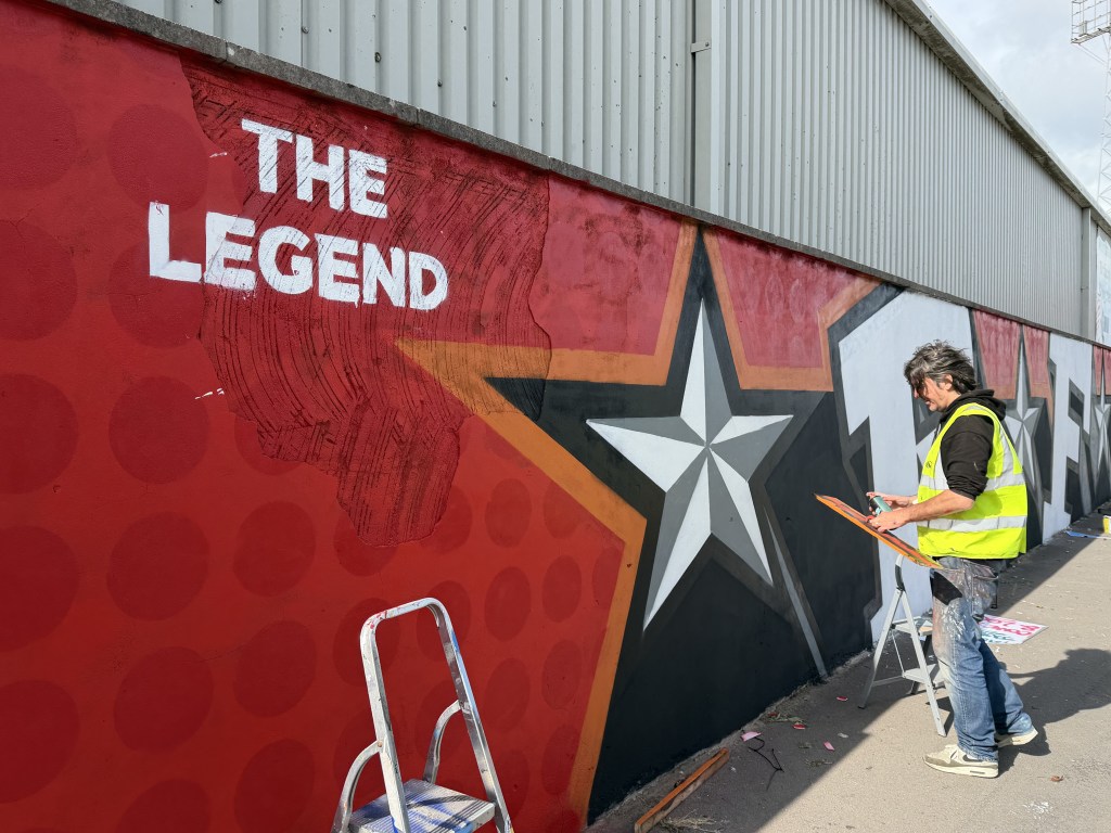 Painter in hi-vis vest working on the mural next to the white stencilled words The Legend on a red background