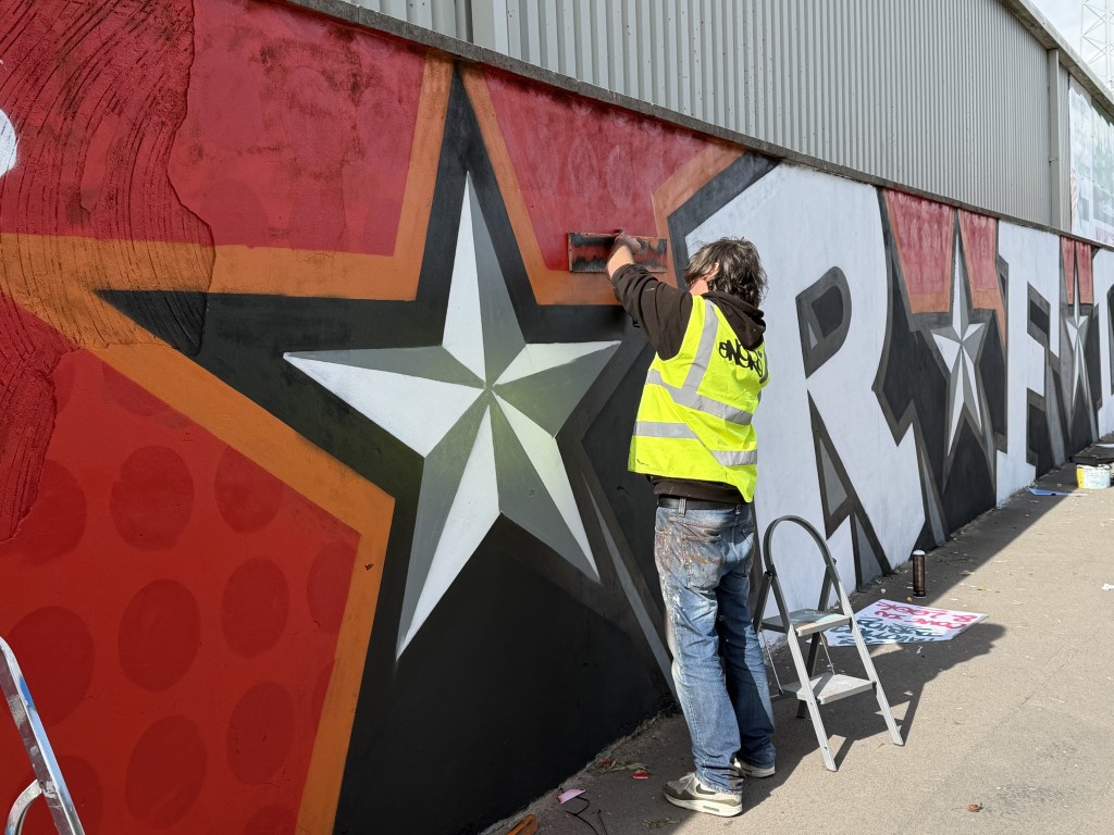 Painter in hi-vis vest using a board to mask an edge while detailing a large silver star on the mural