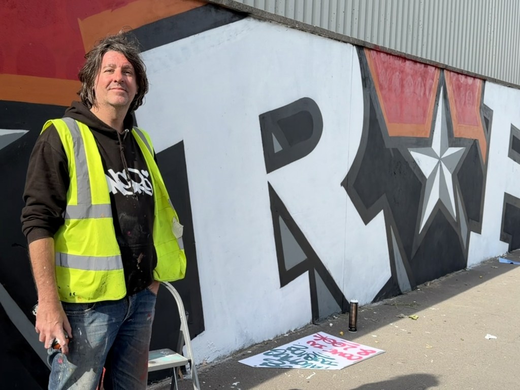 Ronnie Oner in hi-vis vest standing beside the giant letter R and a white star on the St Helen's mural
