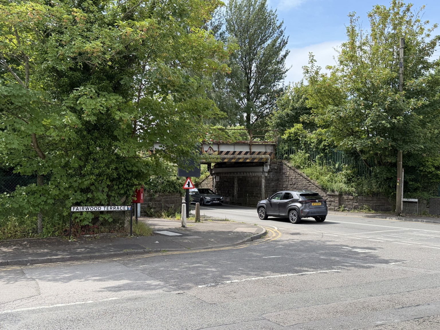 The Fairwood Terrace street sign at its junction with Victoria Road, Gowerton, with a low stone railway bridge crossing the road ahead and traffic passing through — the site of a proposed traffic signal-controlled junction.