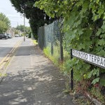 Fairwood Terrace street name sign beside a road leading under a low stone railway bridge, with cars passing.