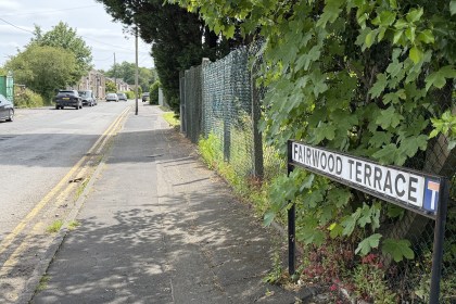 Fairwood Terrace street name sign beside a road leading under a low stone railway bridge, with cars passing.