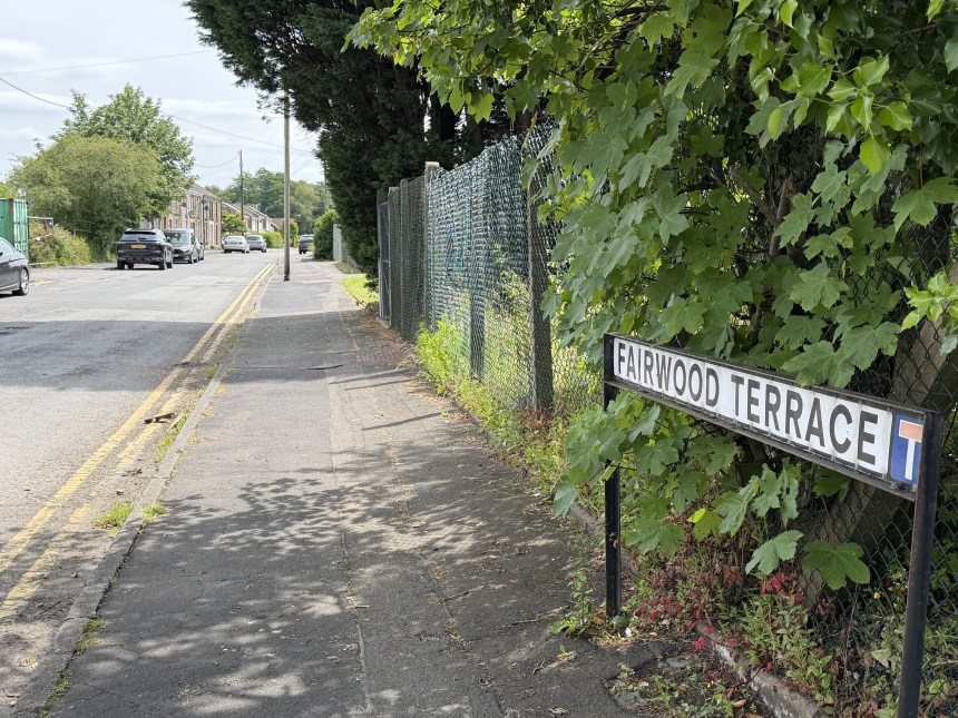 Fairwood Terrace street name sign beside a road leading under a low stone railway bridge, with cars passing.