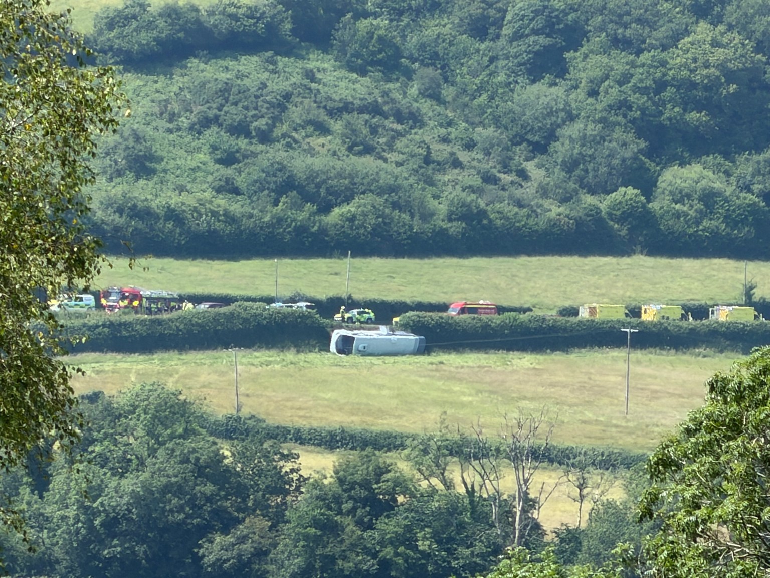 A single-decker bus on its side in a field beside the A484 near Kidwelly, with emergency vehicles including a fire engine on the road behind.