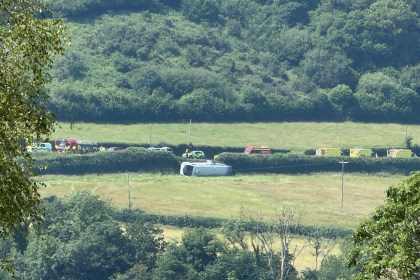 A single-decker bus on its side in a field beside the A484 near Kidwelly, with emergency vehicles including a fire engine on the road behind.