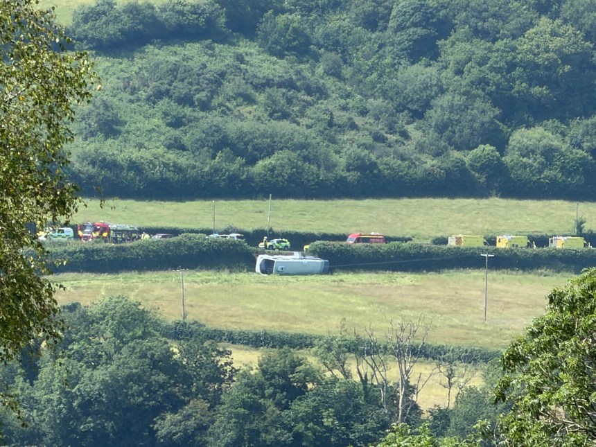 A single-decker bus on its side in a field beside the A484 near Kidwelly, with emergency vehicles including a fire engine on the road behind.