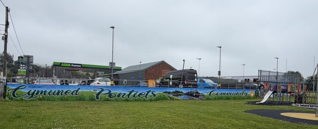 A long painted mural reading "Cymuned Pontiets" along a low wall beside a grassy park and playground.