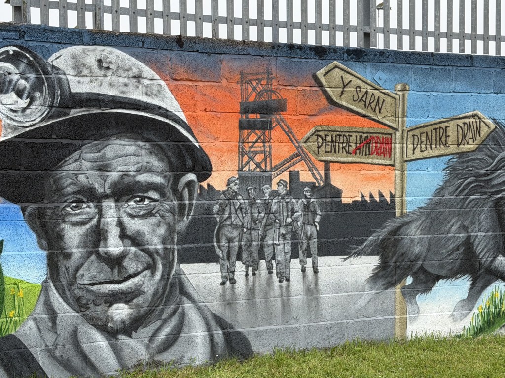 Mural detail of a coal miner in a helmet, a colliery winding tower and a wooden signpost.