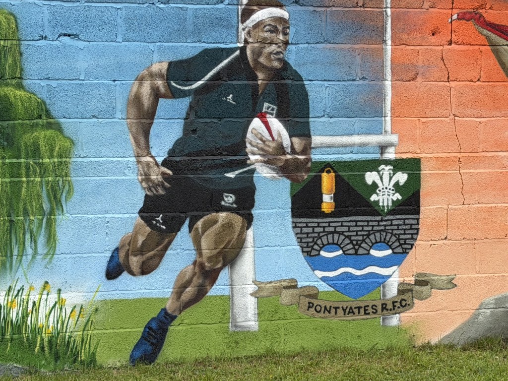 Mural detail of a rugby player in black kit running with the ball beside the Pontyates RFC crest.