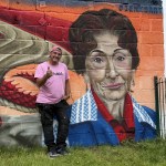 The artist Jenks stands smiling and giving a thumbs-up beside a large painted portrait of Dot Cotton and a Welsh dragon on the mural.