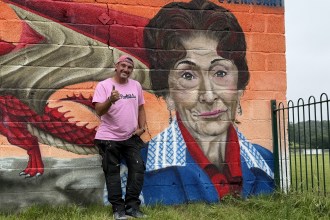 The artist Jenks stands smiling and giving a thumbs-up beside a large painted portrait of Dot Cotton and a Welsh dragon on the mural.