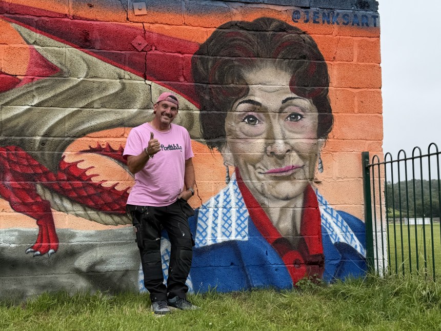 The artist Jenks stands smiling and giving a thumbs-up beside a large painted portrait of Dot Cotton and a Welsh dragon on the mural.