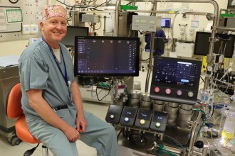 A smiling man in surgical scrubs and a patterned scrub cap sitting beside a heart and lung bypass machine with touchscreen displays in an operating theatre