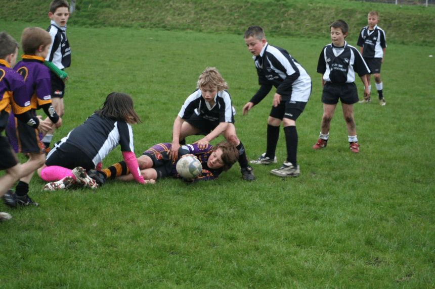 Young children playing junior rugby on a grass pitch, contesting the ball.