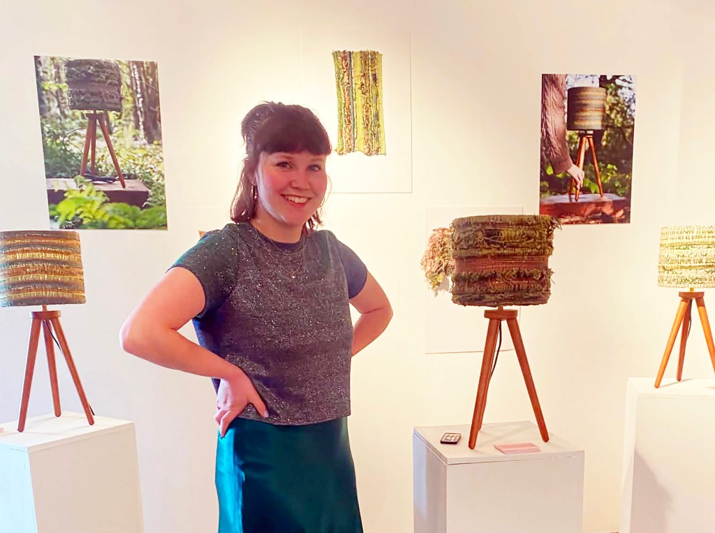 Kitty Haggar smiling in a gallery space surrounded by woven lampshades on wooden tripod stands