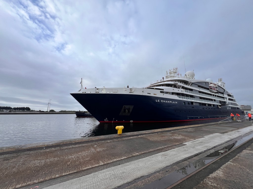 A dark-hulled cruise ship named Le Champlain arriving in port with a tug alongside.
