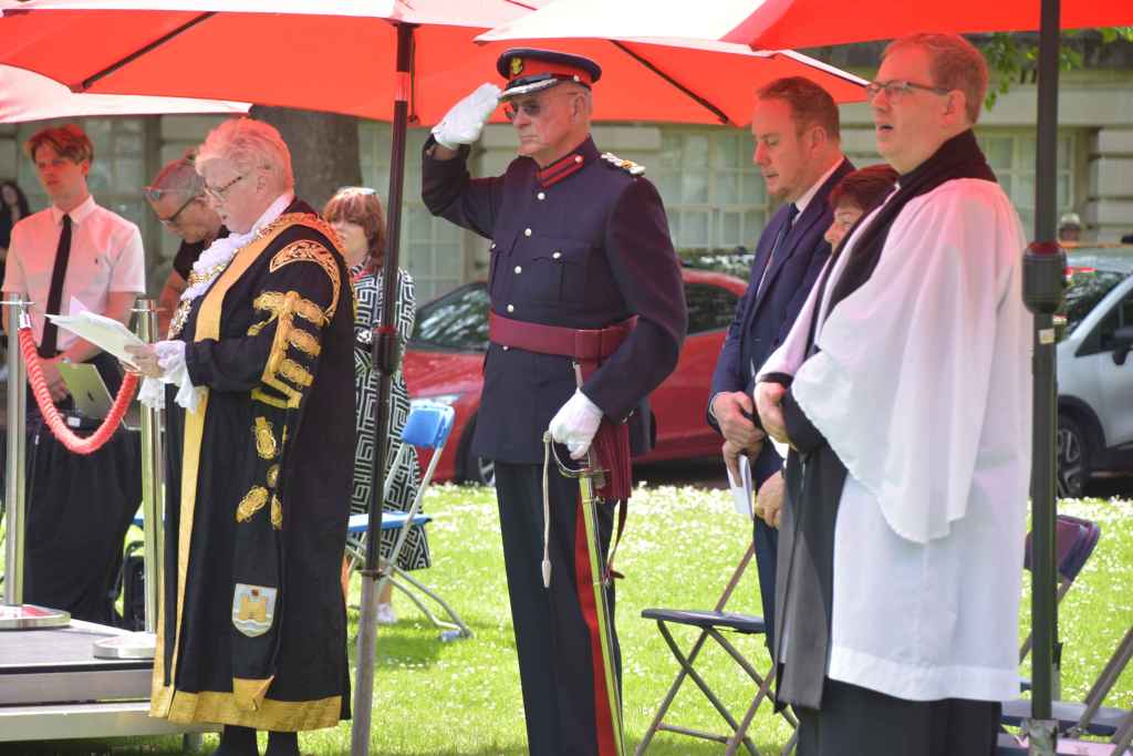 The Lord Mayor of Swansea in ceremonial robes stands beside a uniformed officer saluting, under red parasols at the Guildhall.