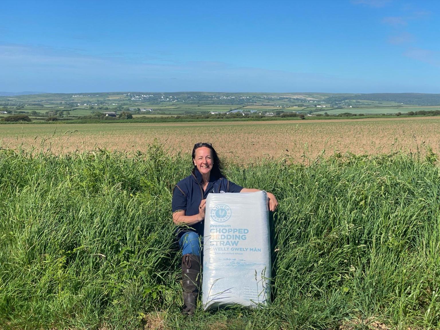A smiling woman kneels in long grass at the edge of a field, holding a large bag labelled "Premium Chopped Bedding Straw", with arable fields, green countryside and a blue sky behind her.