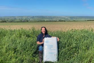 A smiling woman kneels in long grass at the edge of a field, holding a large bag labelled "Premium Chopped Bedding Straw", with arable fields, green countryside and a blue sky behind her.