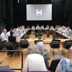 Members of Hywel Dda University Health Board seated around a large table during a public board meeting, with the board's logo on a screen behind them.