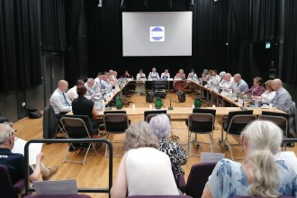 Members of Hywel Dda University Health Board seated around a large table during a public board meeting, with the board's logo on a screen behind them.