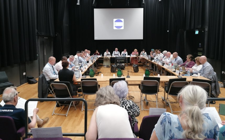 Members of Hywel Dda University Health Board seated around a large table during a public board meeting, with the board's logo on a screen behind them.