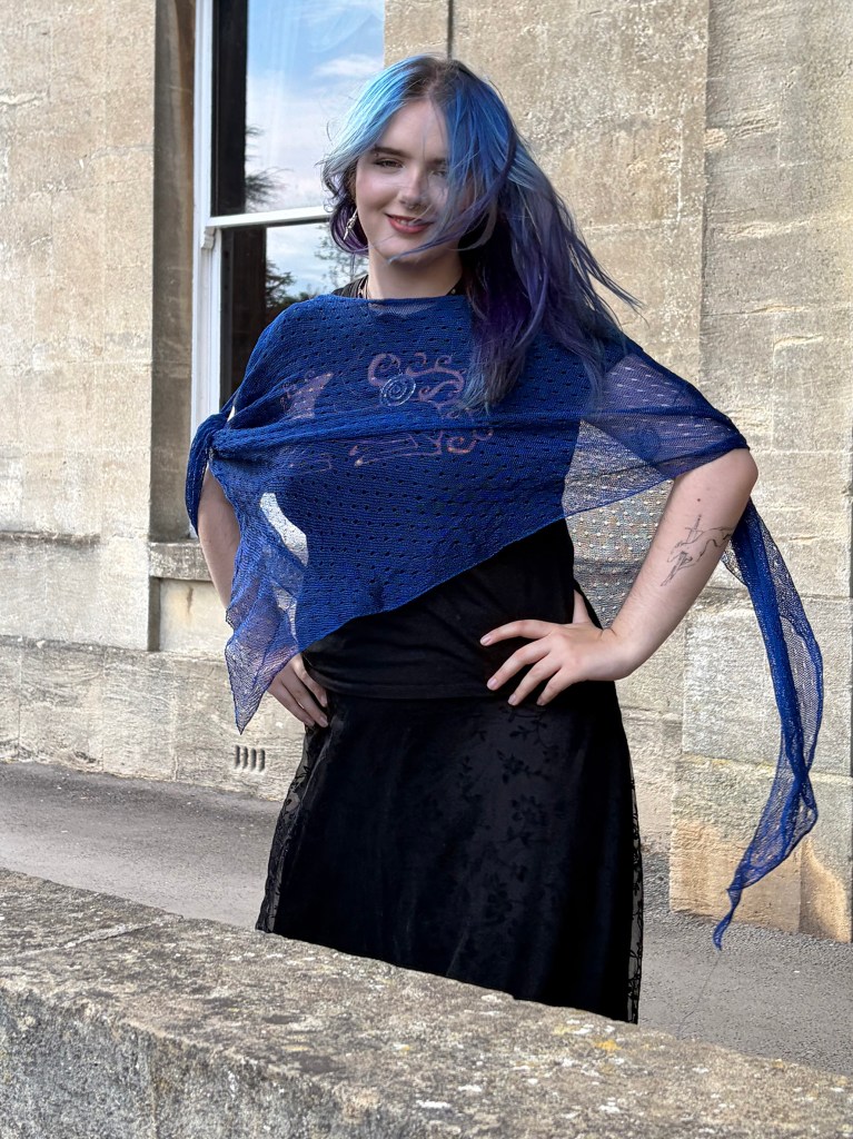 A model wearing a sheer blue knitted shawl draped over a black dress, standing in front of a stone building