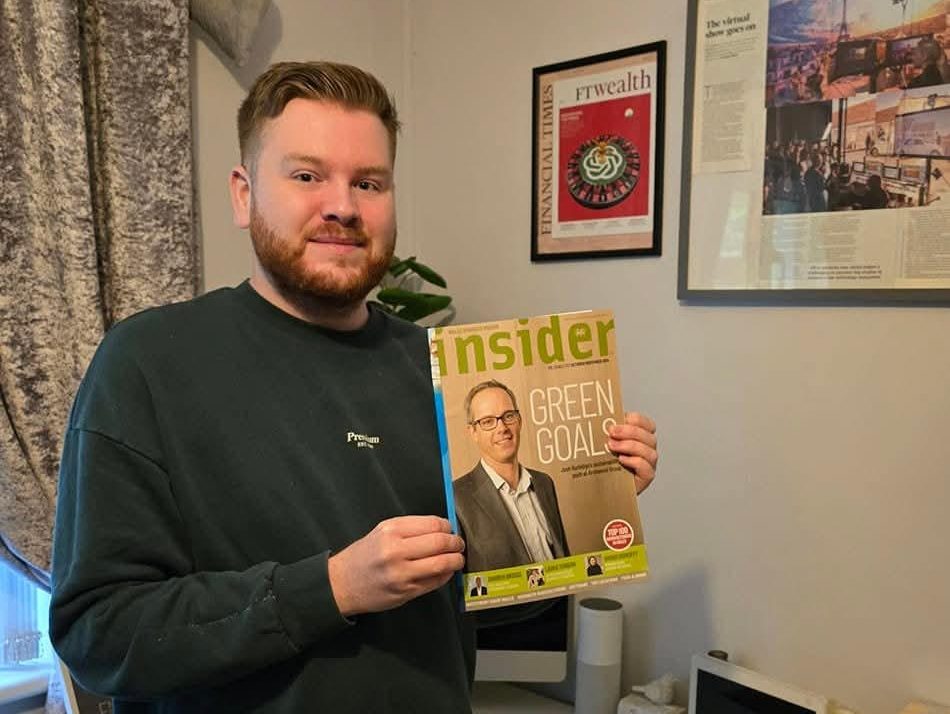 Nicholas Fearn smiling in his home office, holding a copy of Insider business magazine, with framed press cuttings on the wall behind him.