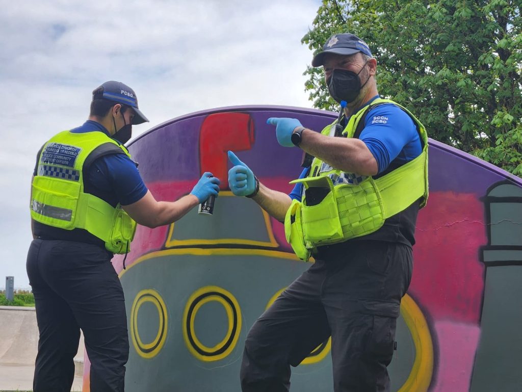 Two police community support officers in hi-vis vests painting a mural, one giving a thumbs-up to camera.
