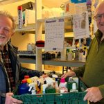 Two men hold a green crate of donated household cleaning products in a food bank storeroom.