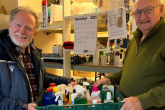 Two men hold a green crate of donated household cleaning products in a food bank storeroom.