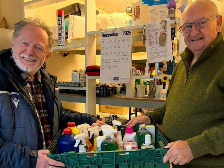 Two men hold a green crate of donated household cleaning products in a food bank storeroom.