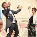 A man in a grey suit throwing his head back mid-shout while a woman in a black dress and white jacket looks on, in a 1960s hotel-room stage set.