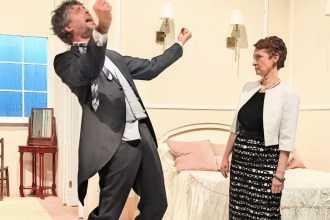 A man in a grey suit throwing his head back mid-shout while a woman in a black dress and white jacket looks on, in a 1960s hotel-room stage set.