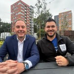 Rob Stewart and Thom Lynch sitting at a bench in a community garden, with city-centre tower blocks behind.