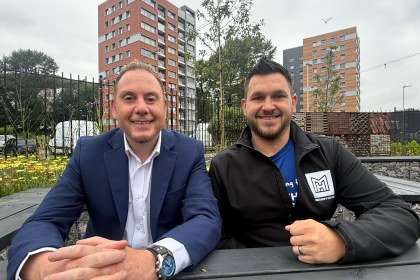 Rob Stewart and Thom Lynch sitting at a bench in a community garden, with city-centre tower blocks behind.
